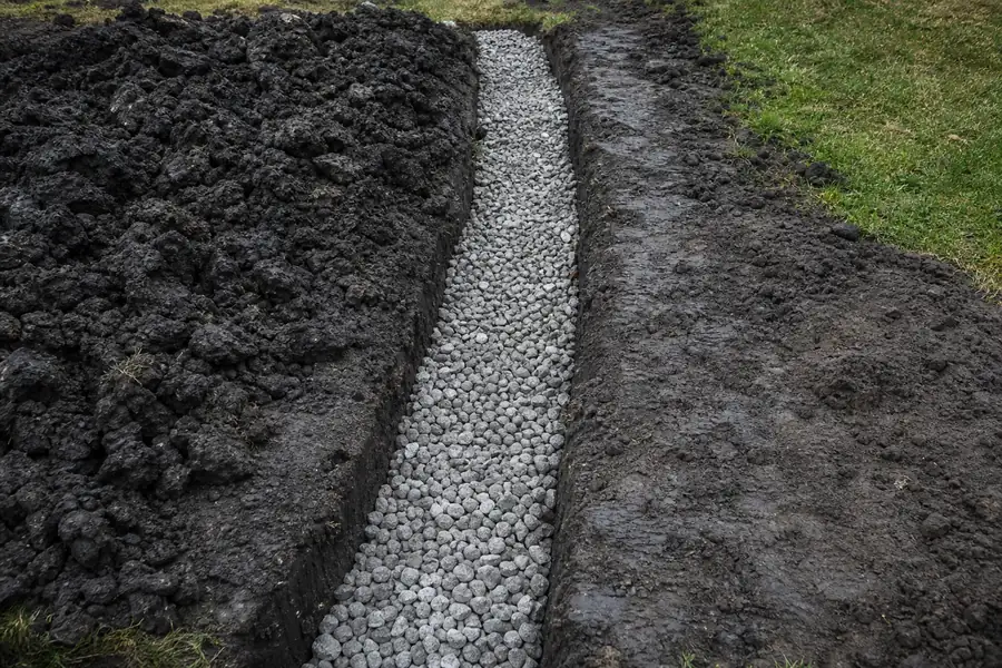French drain trench filled with drain rock cutting through black gumbo clay in a backyard