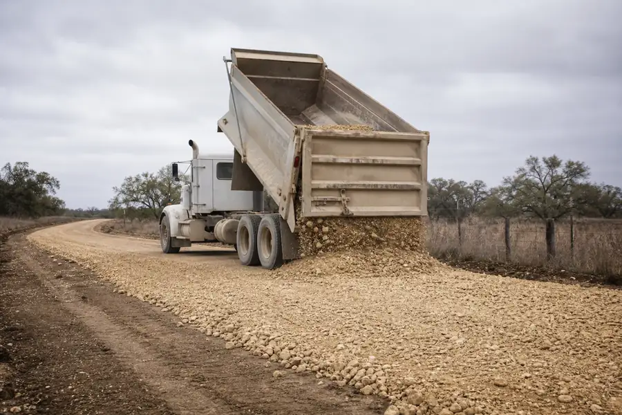 Dump truck spreading caliche base material across a rural Texas ranch road under construction